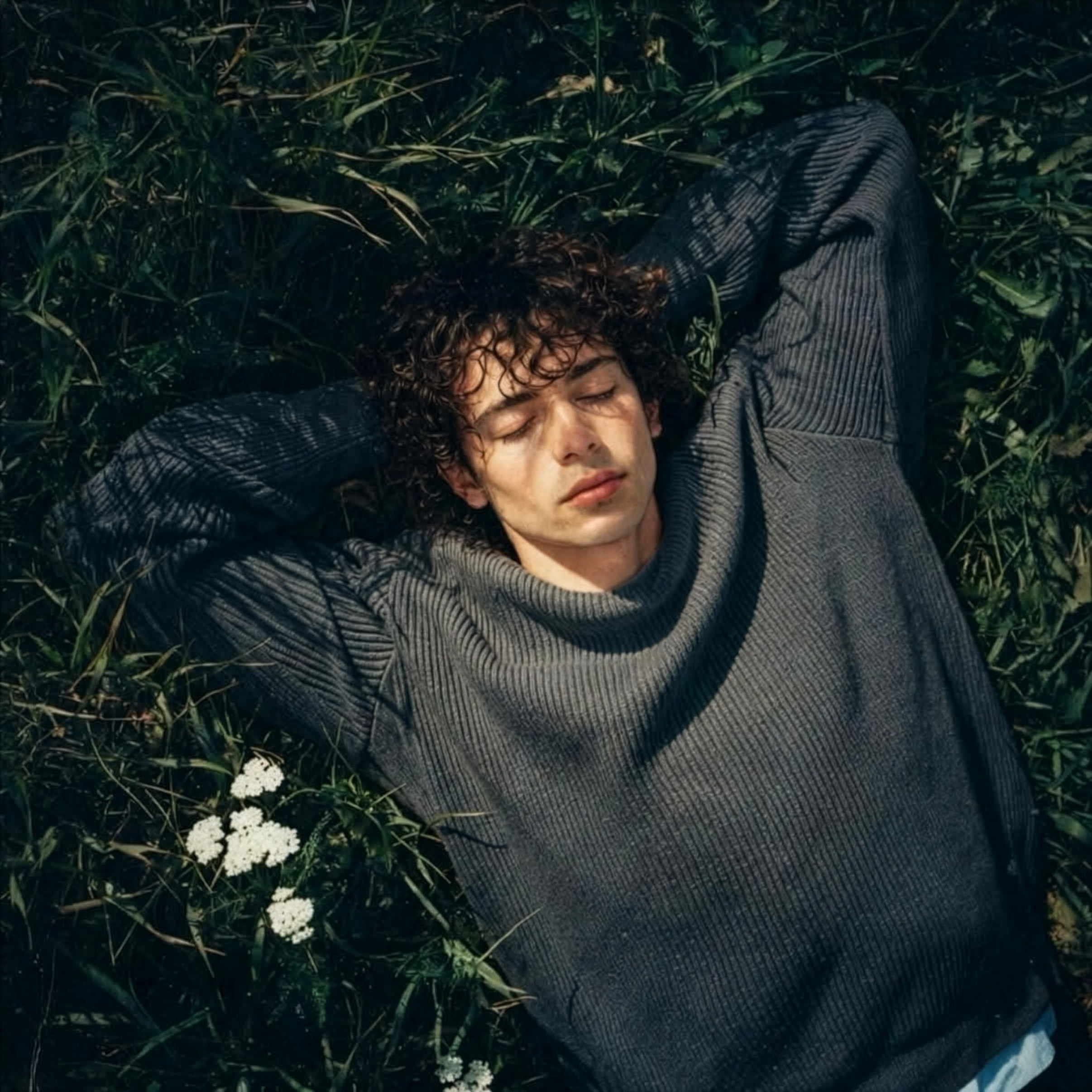 Gage Dunn lying back in dark green grass with eyes closed and hands behind the head, wearing a gray ribbed sweater, white flowers at the lower left